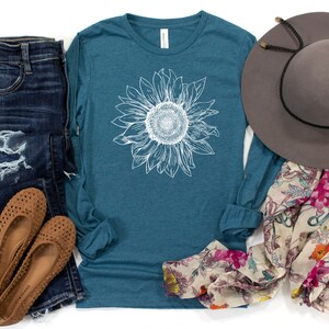 May include: A teal long-sleeve t-shirt with a white sunflower graphic. The shirt is paired with distressed denim jeans, brown sandals, a floral scarf, and a gray fedora.