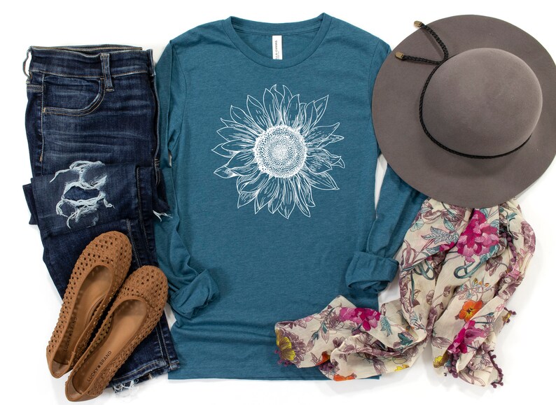 May include: A teal long-sleeve t-shirt with a white sunflower graphic. The shirt is paired with distressed denim jeans, brown sandals, a floral scarf, and a gray fedora.