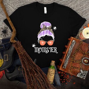 May include: Black t-shirt with a graphic of a woman with a purple bun, leopard print headband, and orange glitter sunglasses. The text "Momster" is printed below the graphic.
