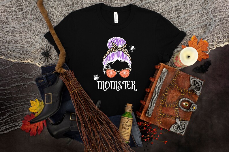 May include: Black t-shirt with a graphic of a woman with a purple bun, leopard print headband, and orange glitter sunglasses. The text "Momster" is printed below the graphic.