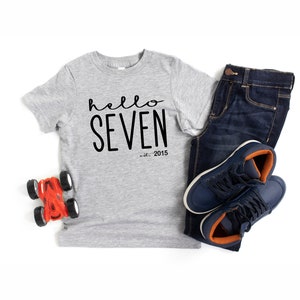 Peut inclure: Un t-shirt gris avec le texte "hello seven est. 2015" en noir. Le t-shirt est sur un fond blanc avec une voiture jouet, un jean bleu et des baskets.
