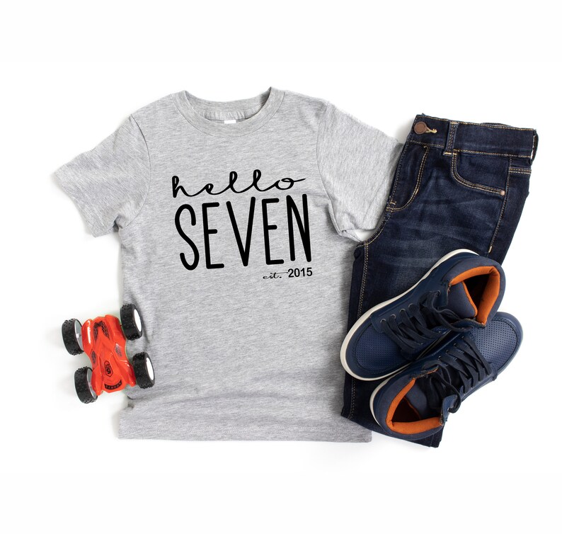 Peut inclure: Un t-shirt gris avec le texte "hello seven est. 2015" en noir. Le t-shirt est sur un fond blanc avec une voiture jouet, un jean bleu et des baskets.