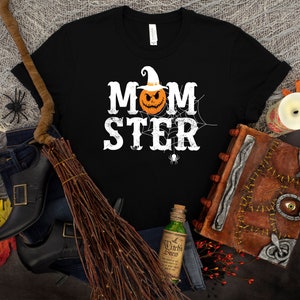 May include: Black t-shirt with a white graphic that says "MOMSTER" with a pumpkin wearing a witch hat in the middle of the word.