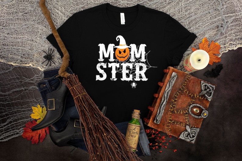 May include: Black t-shirt with a white graphic that says "MOMSTER" with a pumpkin wearing a witch hat in the middle of the word.