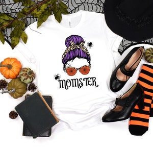 May include: White t-shirt with a graphic of a woman with a purple and leopard print bun, orange spiderweb sunglasses, and the text "Momster".