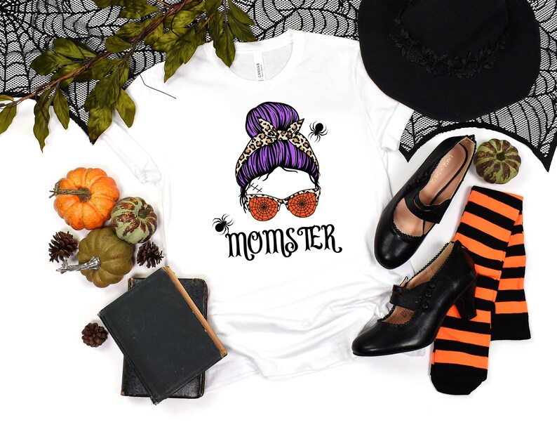 May include: White t-shirt with a graphic of a woman with a purple and leopard print bun, orange spiderweb sunglasses, and the text "Momster".
