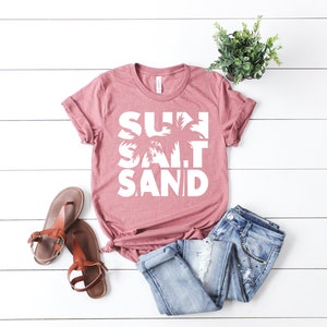 May include: A dusty rose t-shirt with the words "SUN SALT SAND" in white text with a palm tree graphic.