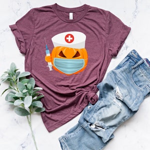 May include: A maroon t-shirt with a graphic of a pumpkin wearing a nurse's hat and a face mask. The pumpkin is holding a syringe.