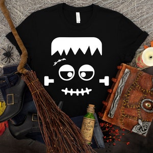 May include: Black t-shirt with a white Frankenstein face graphic. The face has a stitched scar, big eyes, and a stitched mouth.