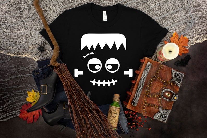 May include: Black t-shirt with a white Frankenstein face graphic. The face has a stitched scar, big eyes, and a stitched mouth.