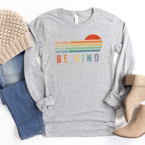 May include: A grey long-sleeved t-shirt with a rainbow stripe design and the text "In a world where you can be anything be kind".