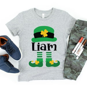 May include: A gray t-shirt with a green leprechaun design, including a hat, legs, and shoes. The name "Liam" is printed in black below the hat.