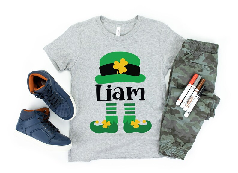 May include: A gray t-shirt with a green leprechaun design, including a hat, legs, and shoes. The name "Liam" is printed in black below the hat.