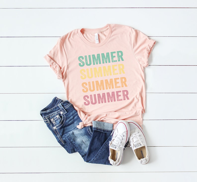 Puede incluir: Una camiseta rosa con la palabra "SUMMER" repetida cuatro veces en un dise&ntilde;o apilado. Cada palabra es de un color diferente: verde, amarillo, naranja y rosa. La camiseta est&aacute; colocada sobre una superficie de madera blanca con un par de pantalones vaqueros azules y zapatillas blancas.