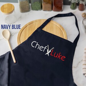 May include: Navy blue apron with the text "Chef Luke" in white and red, featuring crossed knives. A wooden spoon and cutting board are also visible. The apron is designed for kitchen use.