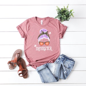May include: A pink t-shirt with a graphic of a woman with a purple messy bun, leopard print headband, and orange spiderweb sunglasses. The text "MOMSTER" is printed below the graphic.