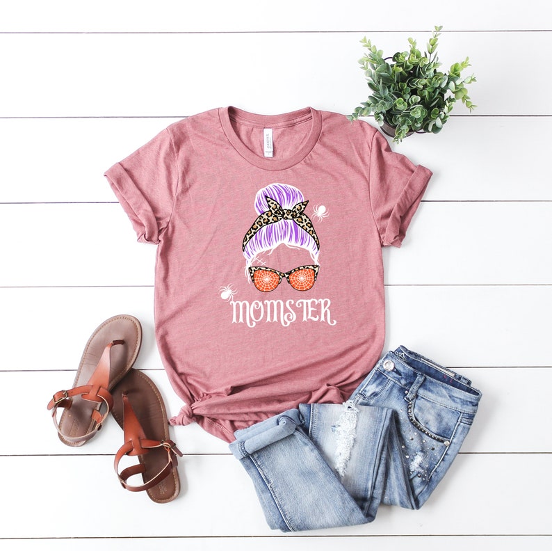 May include: A pink t-shirt with a graphic of a woman with a purple messy bun, leopard print headband, and orange spiderweb sunglasses. The text "MOMSTER" is printed below the graphic.