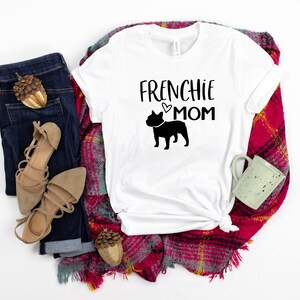 May include: White t-shirt with the text "Frenchie Mom" and a black silhouette of a French Bulldog. The shirt is laid out on a red and white plaid blanket with a pair of brown sandals, blue jeans, and a small brown acorn.