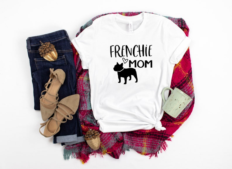 May include: White t-shirt with the text "Frenchie Mom" and a black silhouette of a French Bulldog. The shirt is laid out on a red and white plaid blanket with a pair of brown sandals, blue jeans, and a small brown acorn.