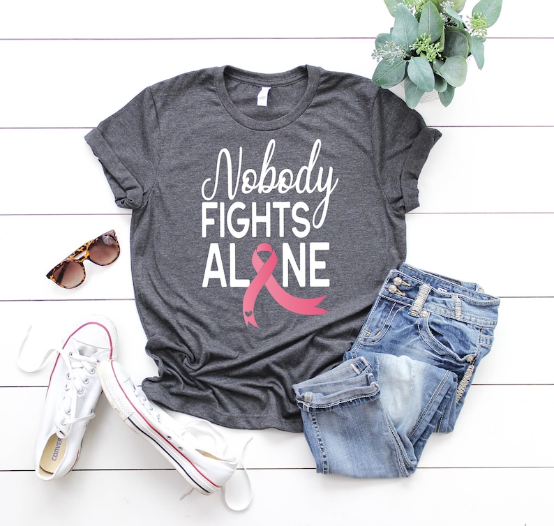May include: A gray t-shirt with the text "Nobody Fights Alone" in white lettering and a pink ribbon with a small heart design.