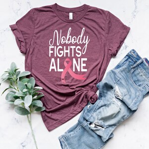 May include: A maroon t-shirt with the text "Nobody Fights Alone" in white lettering and a pink ribbon with two small hearts.