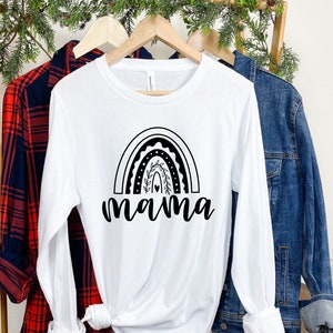 Pu&ograve; includere: Maglietta bianca a maniche lunghe con un design arcobaleno nero e la parola "mama" sotto. La maglietta &egrave; appesa a un appendiabiti di legno con una camicia a quadri rossi e neri e una giacca di jeans blu.