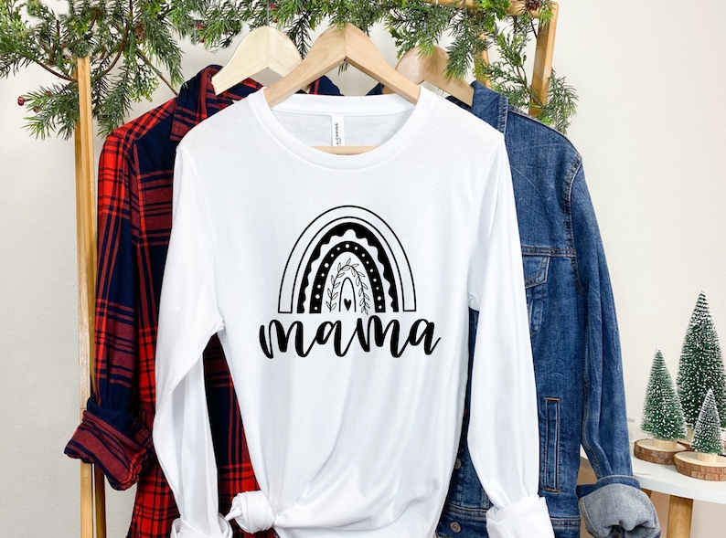 Pu&ograve; includere: Maglietta bianca a maniche lunghe con un design arcobaleno nero e la parola "mama" sotto. La maglietta &egrave; appesa a un appendiabiti di legno con una camicia a quadri rossi e neri e una giacca di jeans blu.