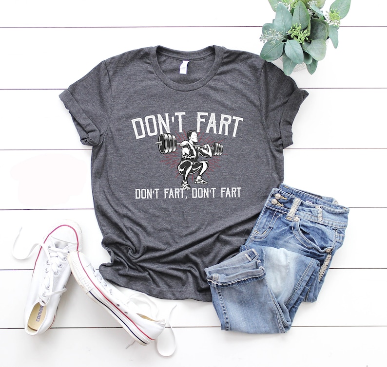 K&ouml;nnte beinhalten: Ein dunkel-helles graues T-Shirt mit einer wei&szlig;en Grafik einer Person, die Gewichte hebt. Der Text "DON'T FART, DON'T FART" ist auf das T-Shirt gedruckt.