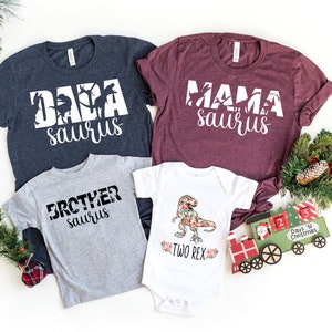 May include: A set of four shirts featuring a dinosaur theme. The shirts are in navy blue, burgundy, grey, and white. The shirts have the text "Dada saurus", "Mama saurus", "Brother saurus", and "Two Rex" printed on them. The shirts are arranged on a white background with a Christmas train and greenery.