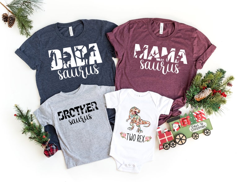 May include: A set of four shirts featuring a dinosaur theme. The shirts are in navy blue, burgundy, grey, and white. The shirts have the text "Dada saurus", "Mama saurus", "Brother saurus", and "Two Rex" printed on them. The shirts are arranged on a white background with a Christmas train and greenery.