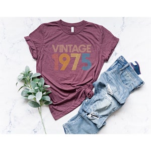 May include: A maroon t-shirt with the text "Vintage 1975" in a rainbow color scheme. The text is in a retro font style.