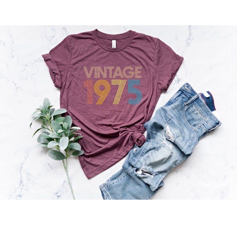 May include: A maroon t-shirt with the text "Vintage 1975" in a rainbow color scheme. The text is in a retro font style.