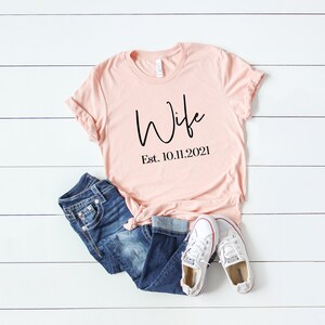 Puede incluir: Una camiseta rosa claro con la palabra "Wife" escrita en cursiva negra y el texto "Est. 10.11.2021" debajo. La camiseta est&aacute; sobre una superficie de madera blanca con un par de pantalones vaqueros azules y zapatillas blancas.