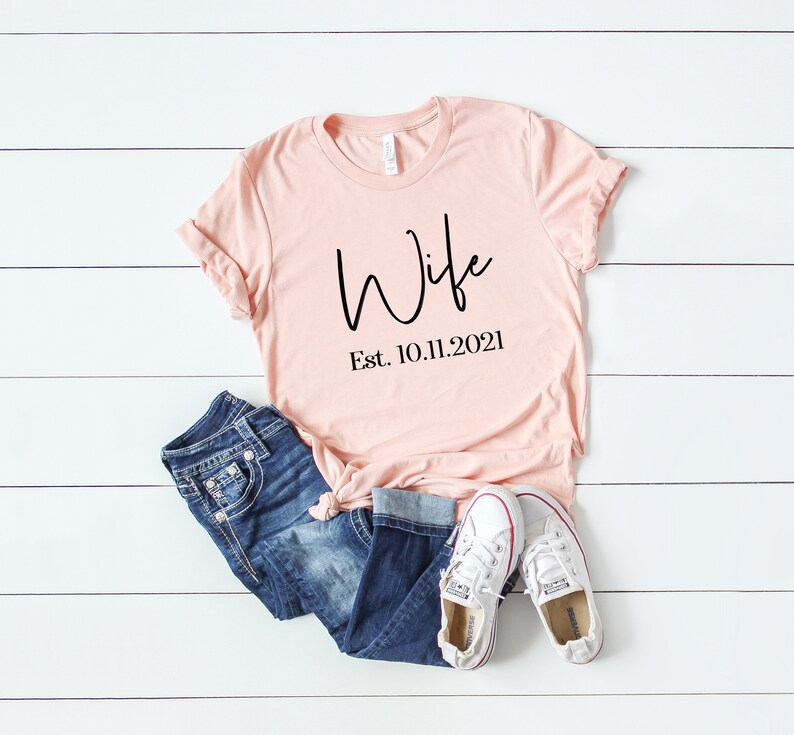 Puede incluir: Una camiseta rosa claro con la palabra "Wife" escrita en cursiva negra y el texto "Est. 10.11.2021" debajo. La camiseta est&aacute; sobre una superficie de madera blanca con un par de pantalones vaqueros azules y zapatillas blancas.