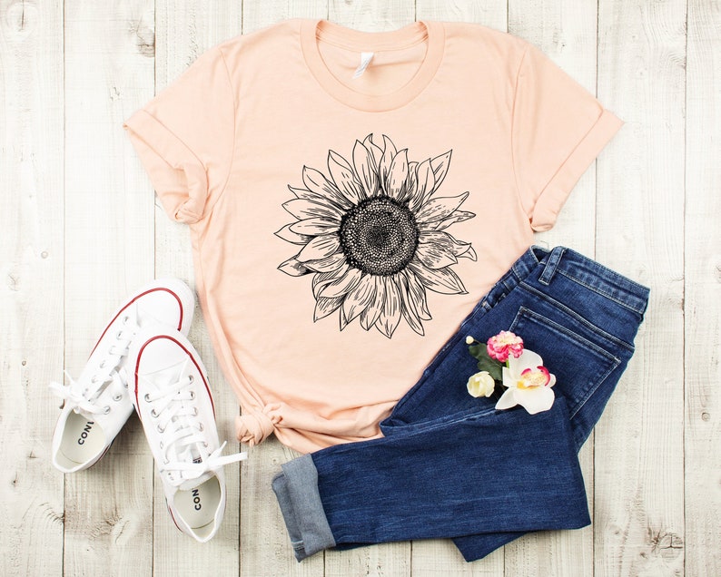 May include: A peach-colored t-shirt with a black and white sunflower graphic. The shirt is laid out on a white wooden surface with a pair of white sneakers and blue jeans.