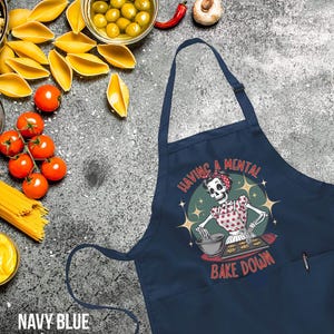 May include: Navy blue apron featuring a skeleton baking cookies. The apron has a pocket on the right side and the text "Having a mental bake down".