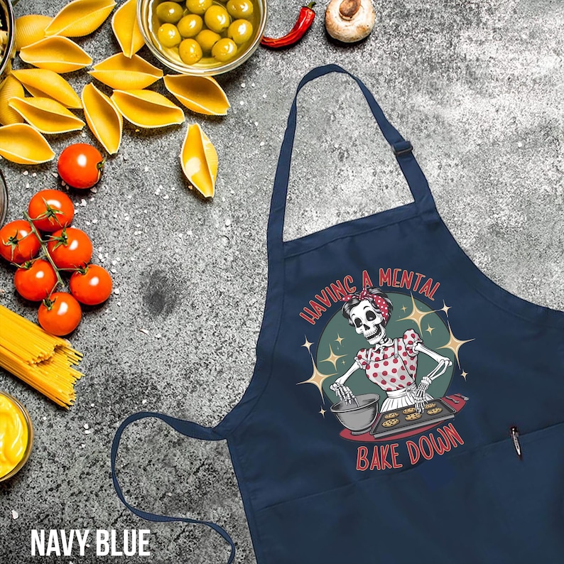 May include: Navy blue apron featuring a skeleton baking cookies. The apron has a pocket on the right side and the text "Having a mental bake down".