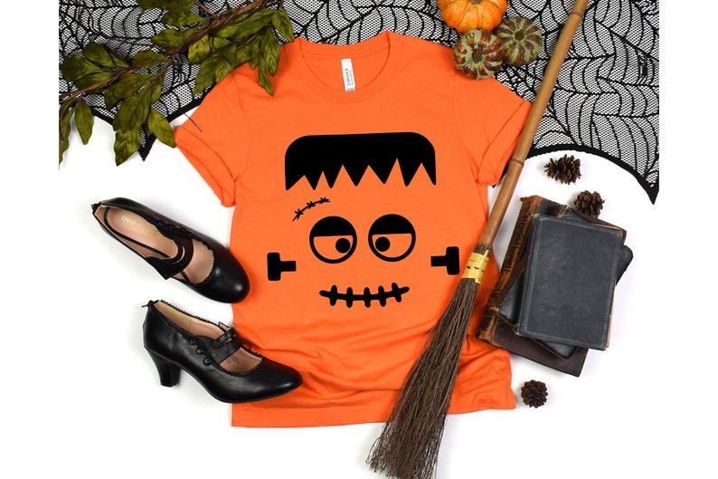 May include: Orange t-shirt with a black Frankenstein face graphic. The shirt is laying flat on a white background with a broom, books, and a black spiderweb.