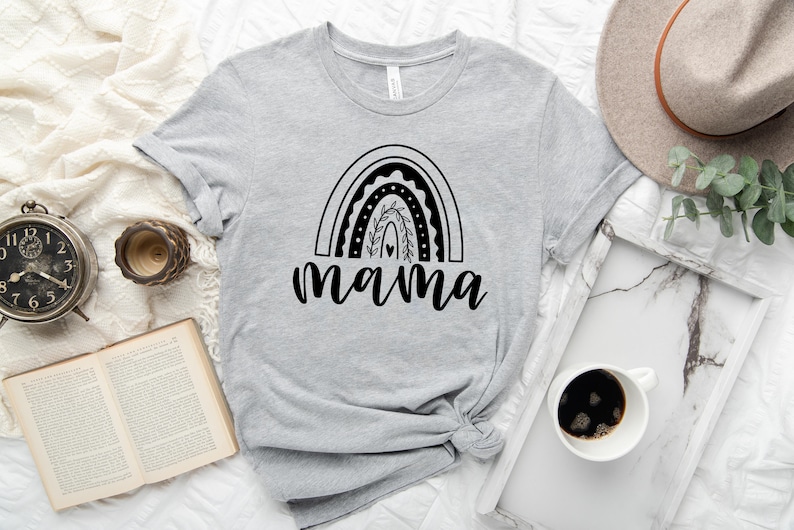 Pu&ograve; includere: Una maglietta grigia con un design arcobaleno nero e la parola "mamma" sotto. L'arcobaleno ha un cuore e foglie al centro.