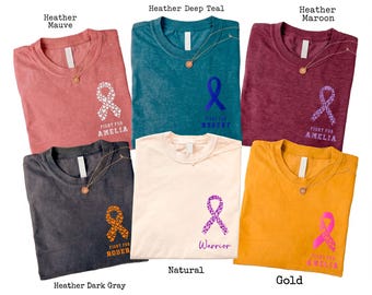 May include: A collection of six t-shirts in various colors, including heather mauve, deep teal, maroon, dark gray, natural, and gold. Each shirt features a ribbon graphic and text, such as "Fight for Amelia" and "Warrior."