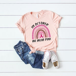 May include: A pink t-shirt with a rainbow design and the text "In October We Wear Pink". The rainbow is made of pink, brown, and white glitter and has a pink ribbon in the center.