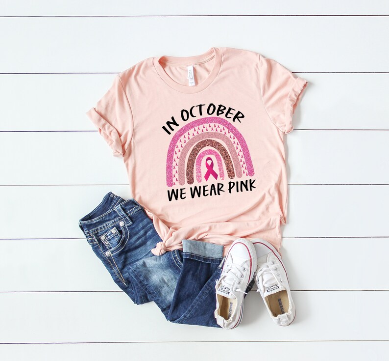 May include: A pink t-shirt with a rainbow design and the text "In October We Wear Pink". The rainbow is made of pink, brown, and white glitter and has a pink ribbon in the center.