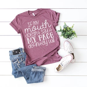 May include: A maroon t-shirt with the slogan "If my mouth doesn't say it, my face definitely will" in white. The image includes blue jeans and white Converse trainers. A small potted plant is also present.
