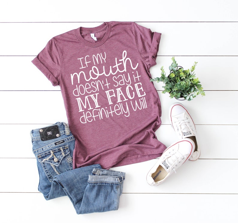 May include: A maroon t-shirt with the slogan "If my mouth doesn't say it, my face definitely will" in white. The image includes blue jeans and white Converse trainers. A small potted plant is also present.