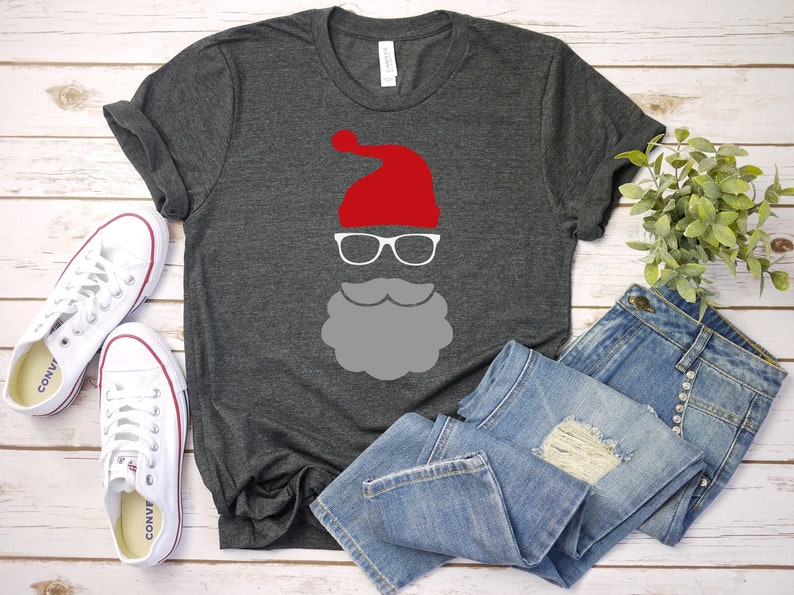 May include: A gray t-shirt with a white Santa Claus face design. The design includes a red hat, white glasses, and a gray beard.