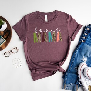 May include: A maroon t-shirt with the text "bonus mama" in white lettering. The letters are decorated with colorful patterns and leopard print.