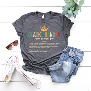 May include: A gray t-shirt with a colorful graphic design that reads "Black Grandpa" with a definition and a crown above the word. The definition reads "A man who has stepped up to the challenge of raising his children to be Kings and Queens. Someone who has risen above the negative stereotypes about Black Fathers and is a King in his own right. A provider, a protector, a lover and a strong foundation. Royalty in his lineage."