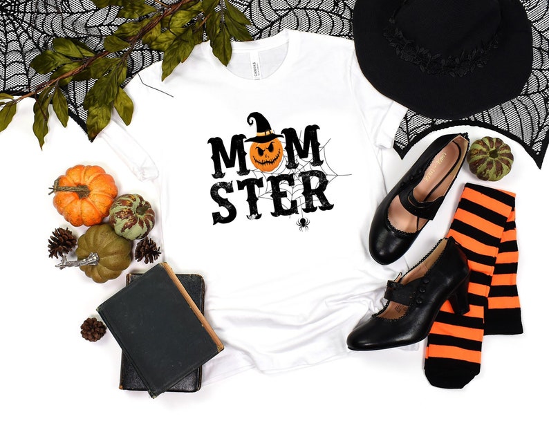 May include: White t-shirt with a black and orange pumpkin graphic and the text "MOMSTER" in black. The shirt is surrounded by Halloween decorations, including a witch's hat, black shoes, and orange and green pumpkins.