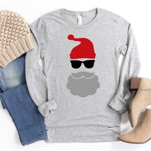 May include: A gray long-sleeved shirt with a red Santa hat and black sunglasses graphic. The shirt is paired with blue jeans and brown ankle boots.