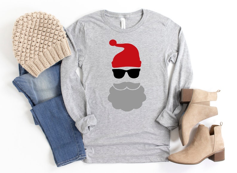 May include: A gray long-sleeved shirt with a red Santa hat and black sunglasses graphic. The shirt is paired with blue jeans and brown ankle boots.